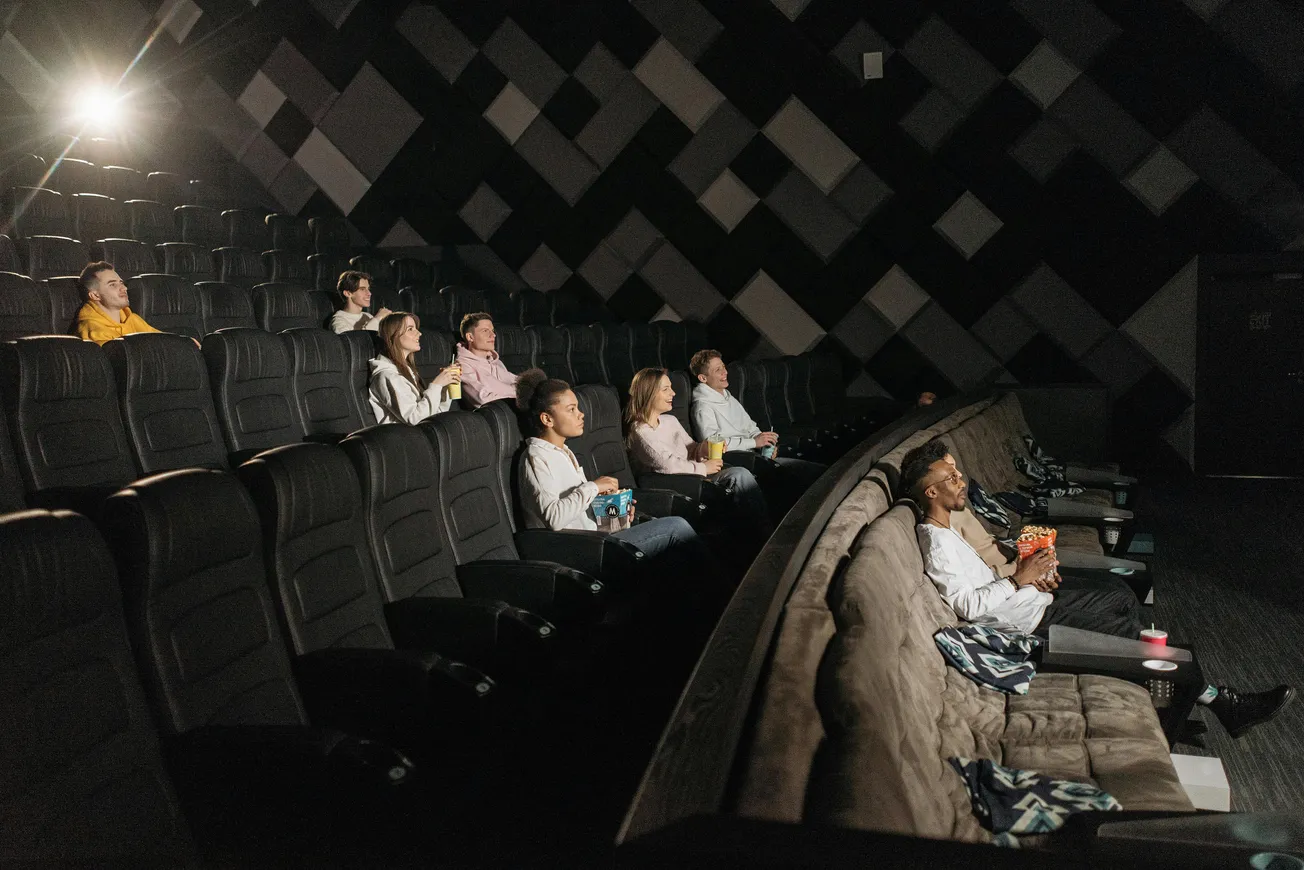 Seven people are sitting in a dimly-lit movie theater, spaced out in plush seats. They appear absorbed, with snacks in hand, creating a cozy atmosphere.