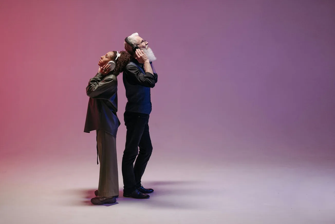 Two people stand back-to-back, wearing headphones, immersed in music. The background features a gradient of pink and purple, creating a relaxed mood.