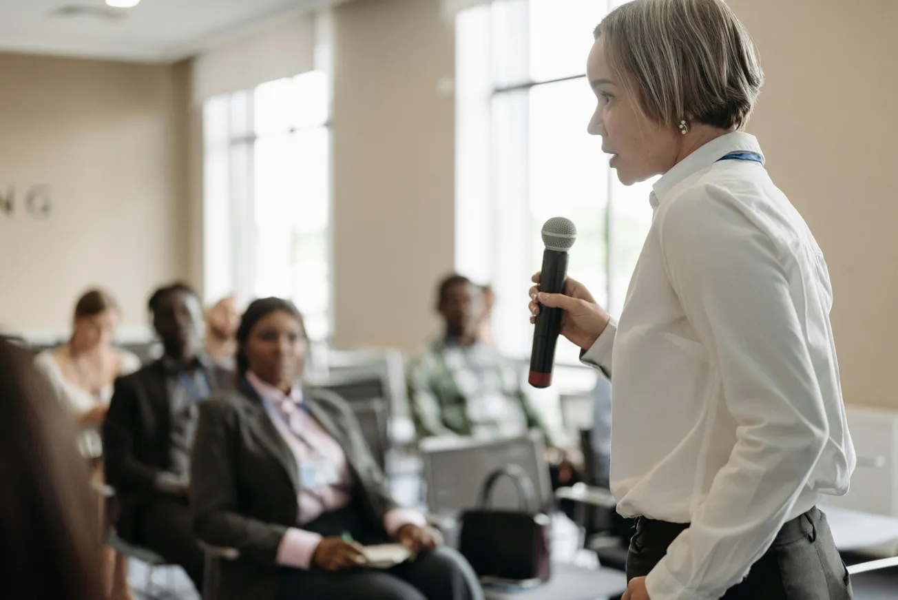 A person in a white shirt is speaking into a microphone, addressing an attentive audience in a conference room.