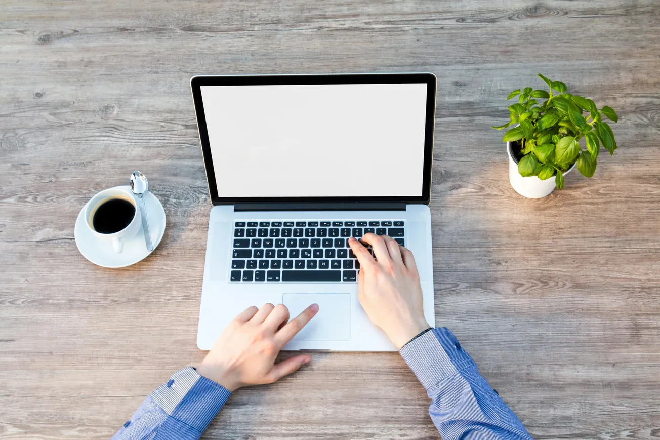 Hands typing on a laptop with a blank screen on a wooden desk. A coffee cup and a potted plant are placed on either side, creating a focused work atmosphere.