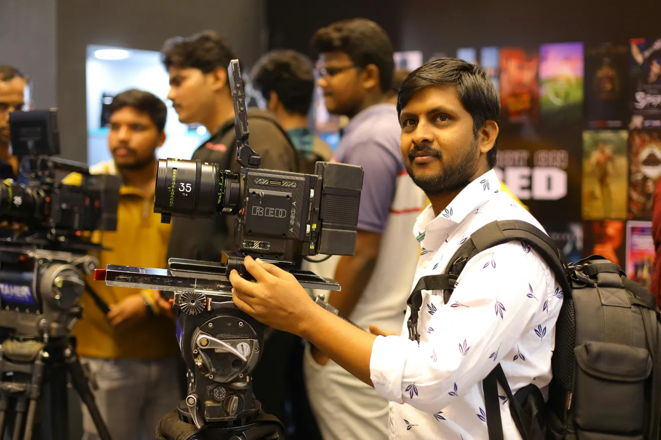 A man with a backpack stands smiling and operating a professional camera on a tripod at an event. Several people and colorful posters are in the background.