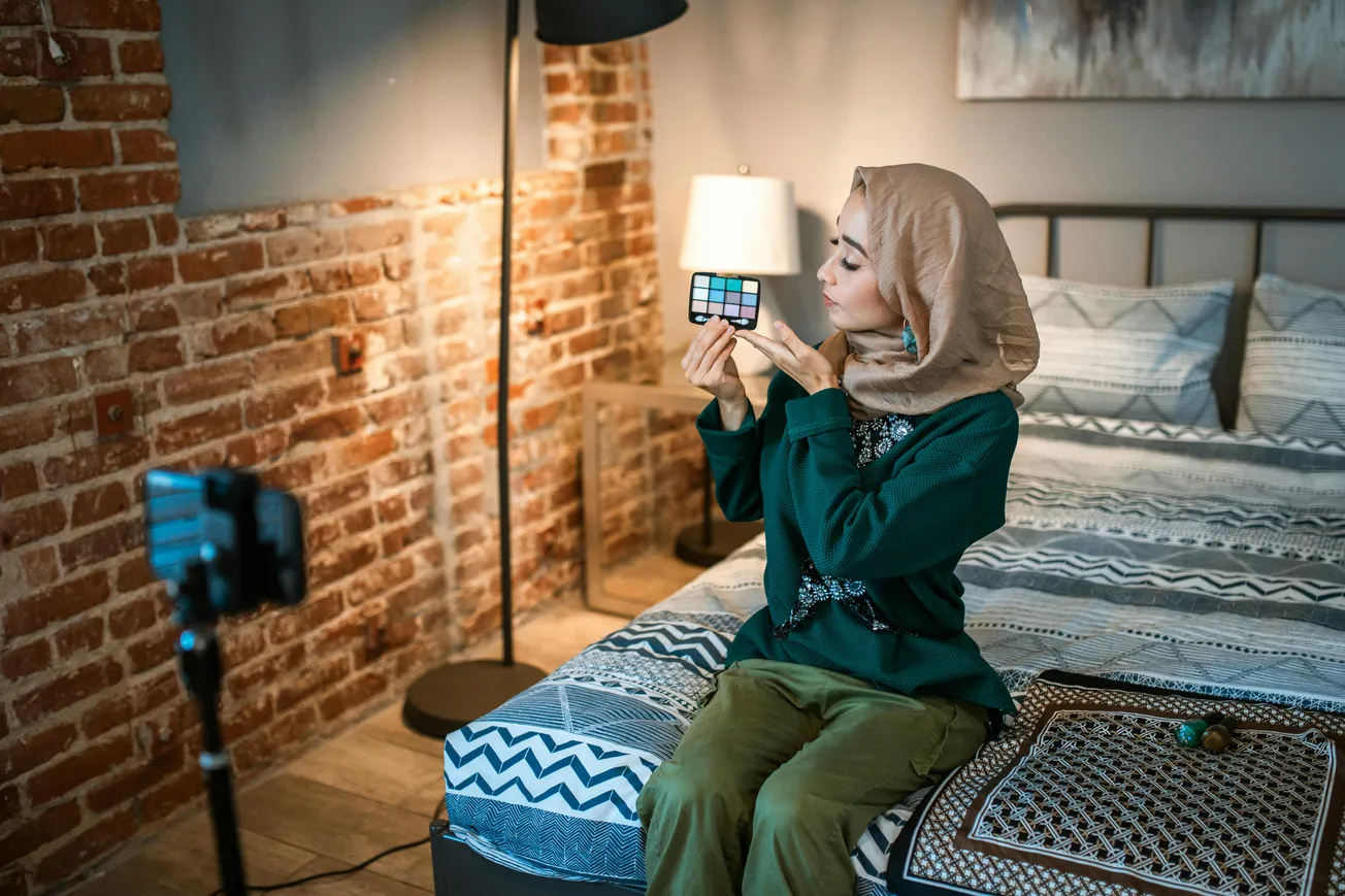 A woman in a headscarf sits on a bed in a cozy, brick-walled room, holding a makeup palette. A camera on a tripod records her tutorial. The atmosphere is calm and focused.