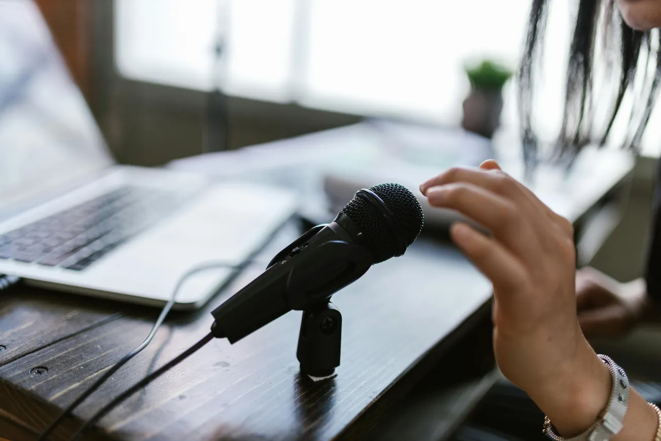 A person adjusts a microphone on a wooden desk beside a laptop, suggesting a podcast or recording setting. The atmosphere is focused and professional.