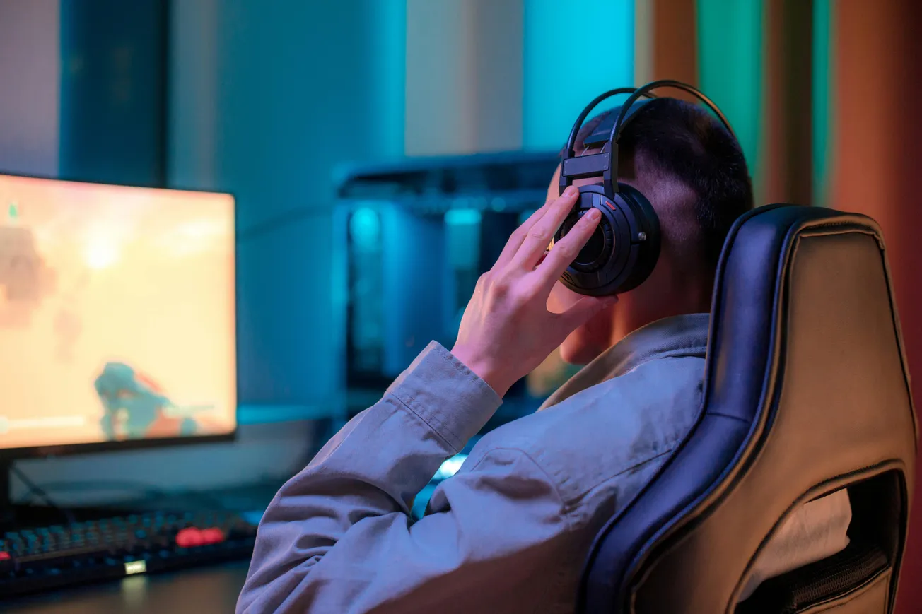 A person wearing headphones sits in a gaming chair, focused on a glowing monitor. The room is lit with blue and orange hues, creating an immersive gaming ambiance.