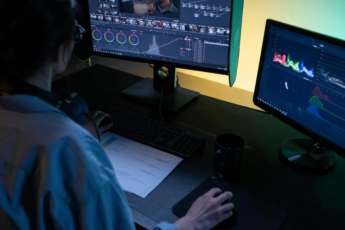 A person edits footage on dual monitors, displaying color grading tools and timelines. The dimly lit room creates a focused, professional atmosphere.