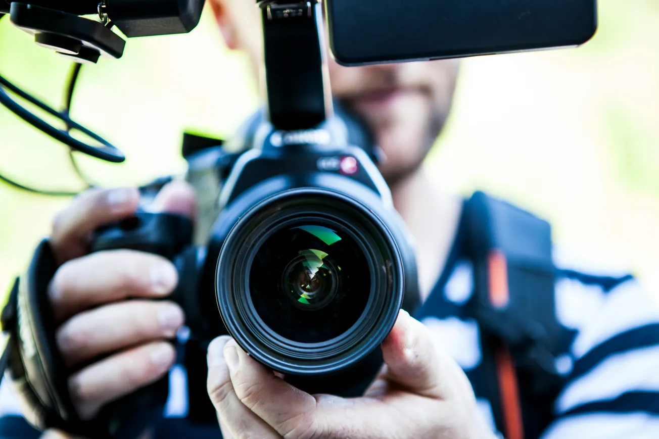 Close-up of a person holding a professional camera with a visible lens and microphone. The background is softly blurred.