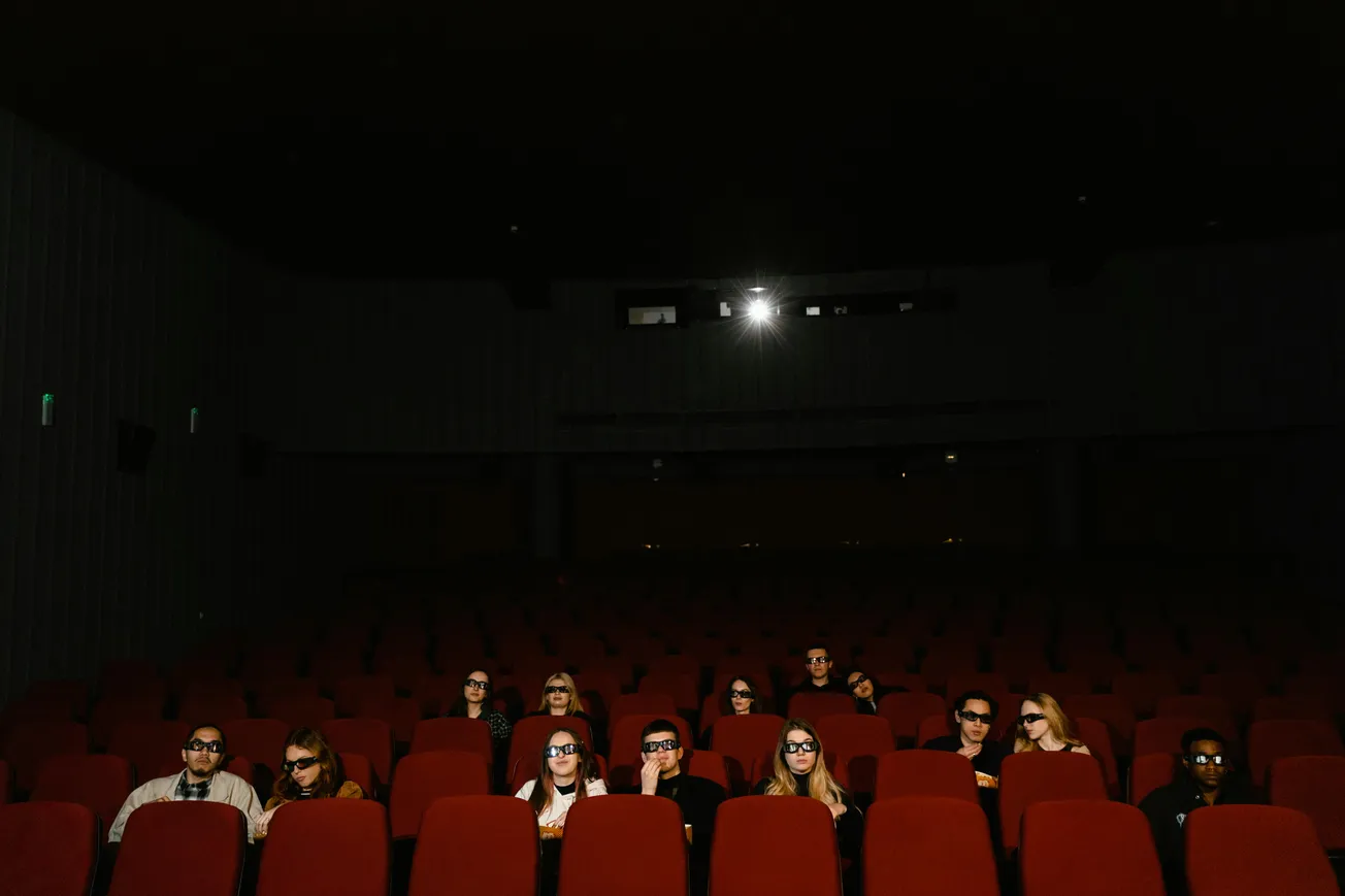 A dimly lit cinema with scattered audience members wearing 3D glasses. They sit in red seats, watching a film with a focused expression.