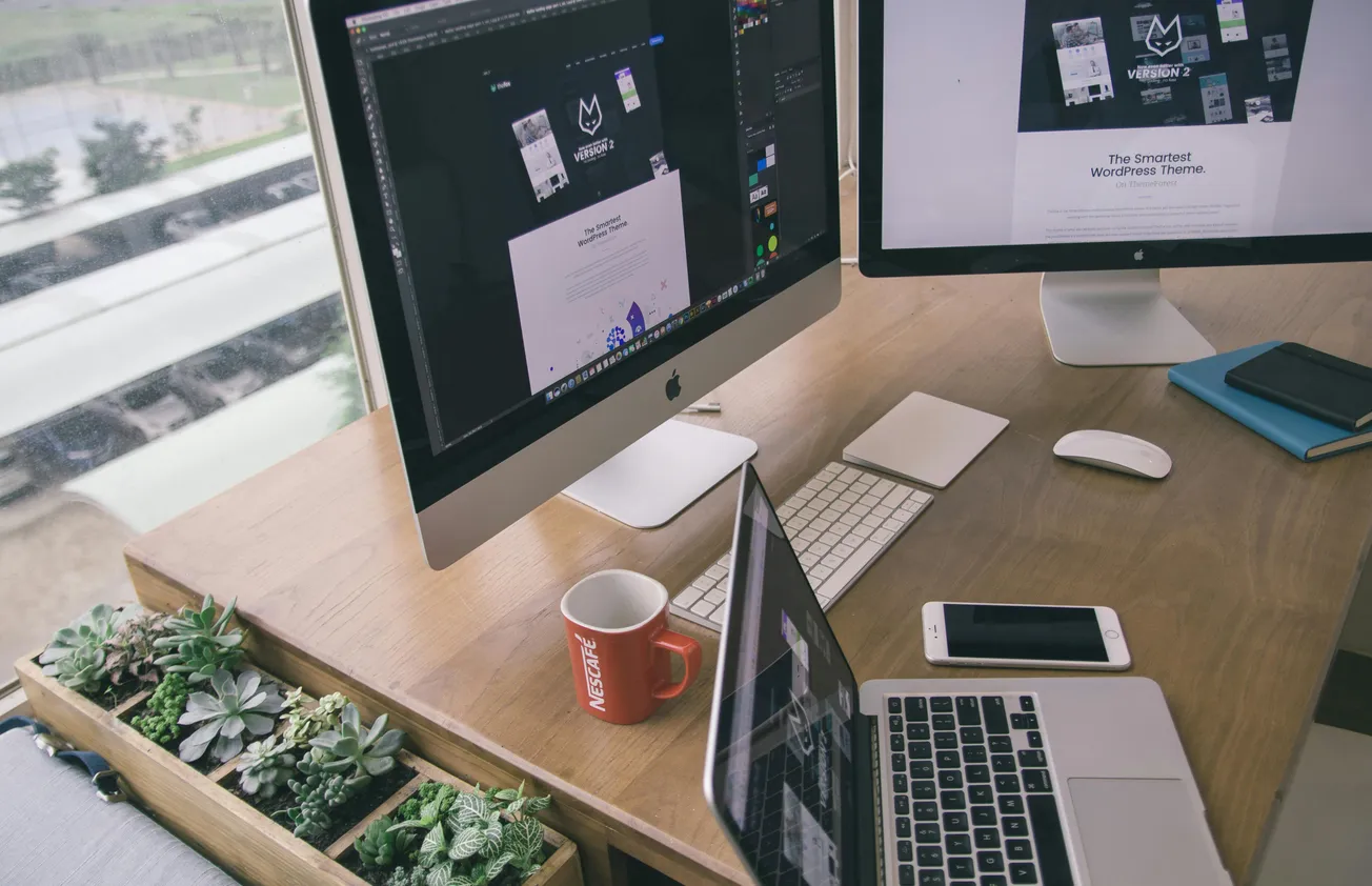 Modern office desk with two monitors displaying design software, a laptop, phone, red coffee mug, notebooks, and a planter with succulents.