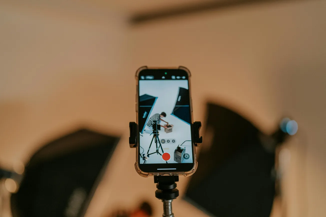 A smartphone on a tripod records a studio setup, showing softbox lights and a person adjusting equipment. The setting conveys a professional, creative tone.