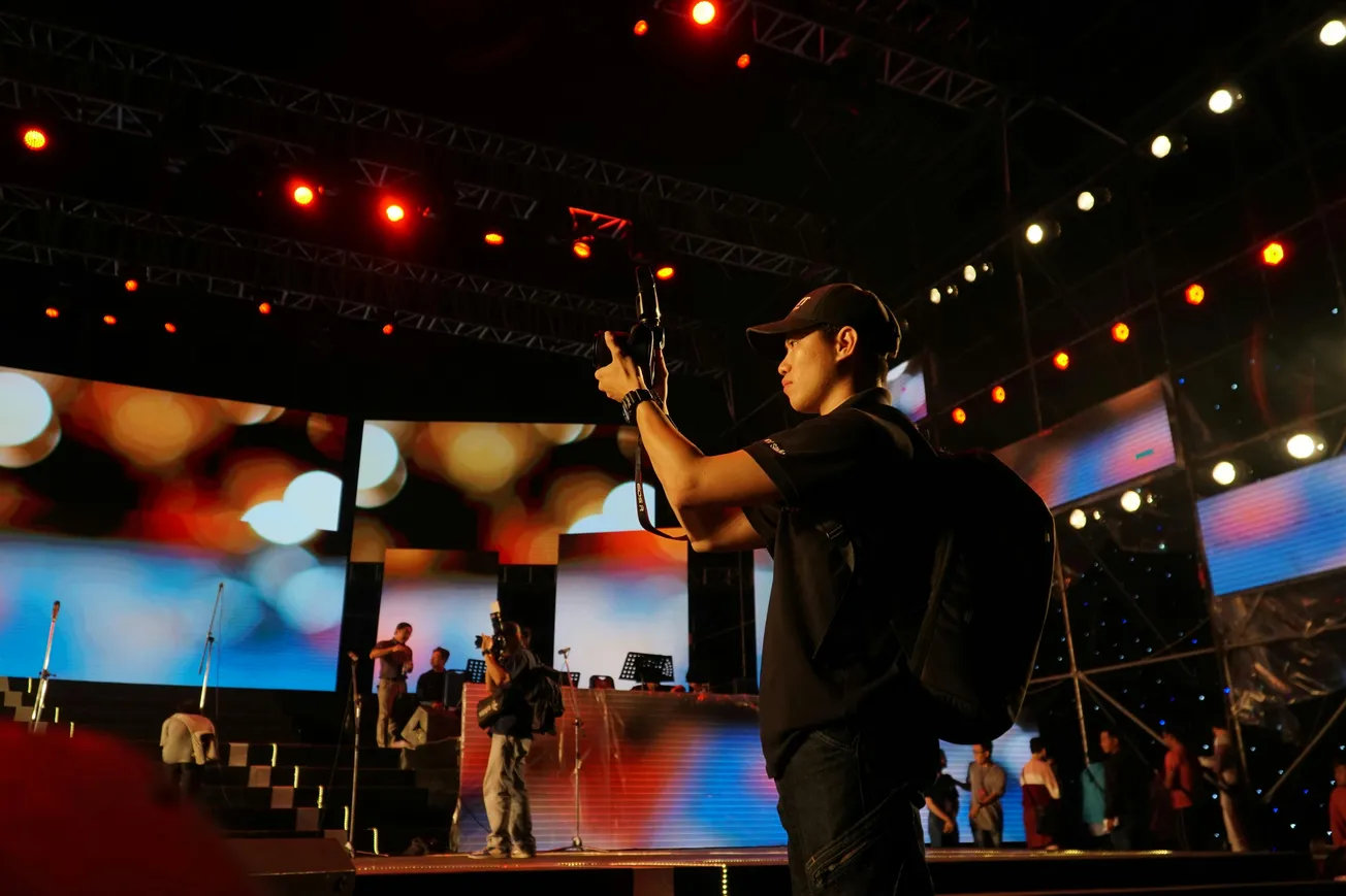 A photographer with a backpack stands on a vibrant stage, capturing the scene. Bright lights and colorful screens create an energetic, dynamic atmosphere.