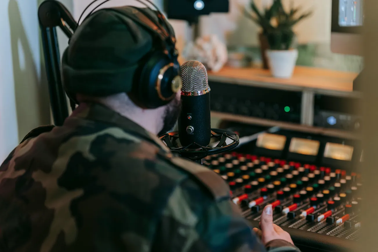 Man in camouflage jacket and headphones recording in a studio, using a microphone and audio mixing console. Cozy ambiance with a plant in the background.