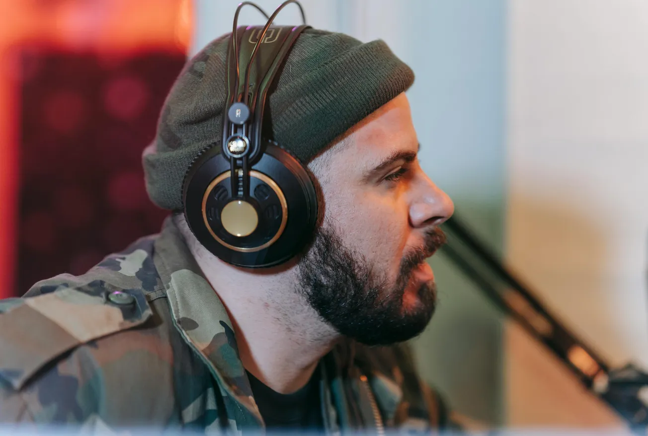 A man with a beard in a beanie and camouflage jacket wears large headphones and speaks into a microphone in a studio setting, focused and engaged.