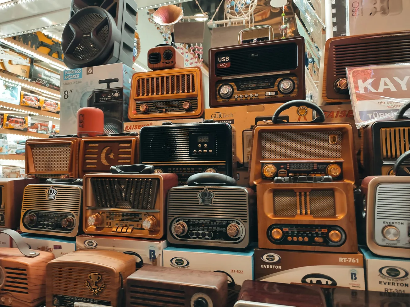 A colorful display of vintage radios stacked in a store, showcasing various styles and designs. Warm lighting highlights their retro appeal.