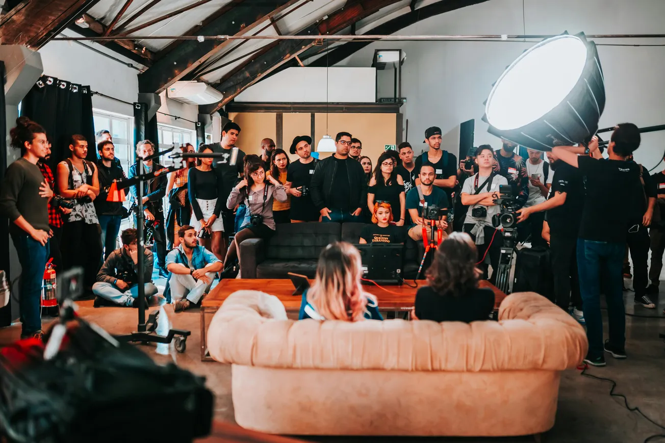 A large group of people gathers in an industrial studio with high ceilings, focused on a bright light setup. Two people sit on a beige sofa observing.
