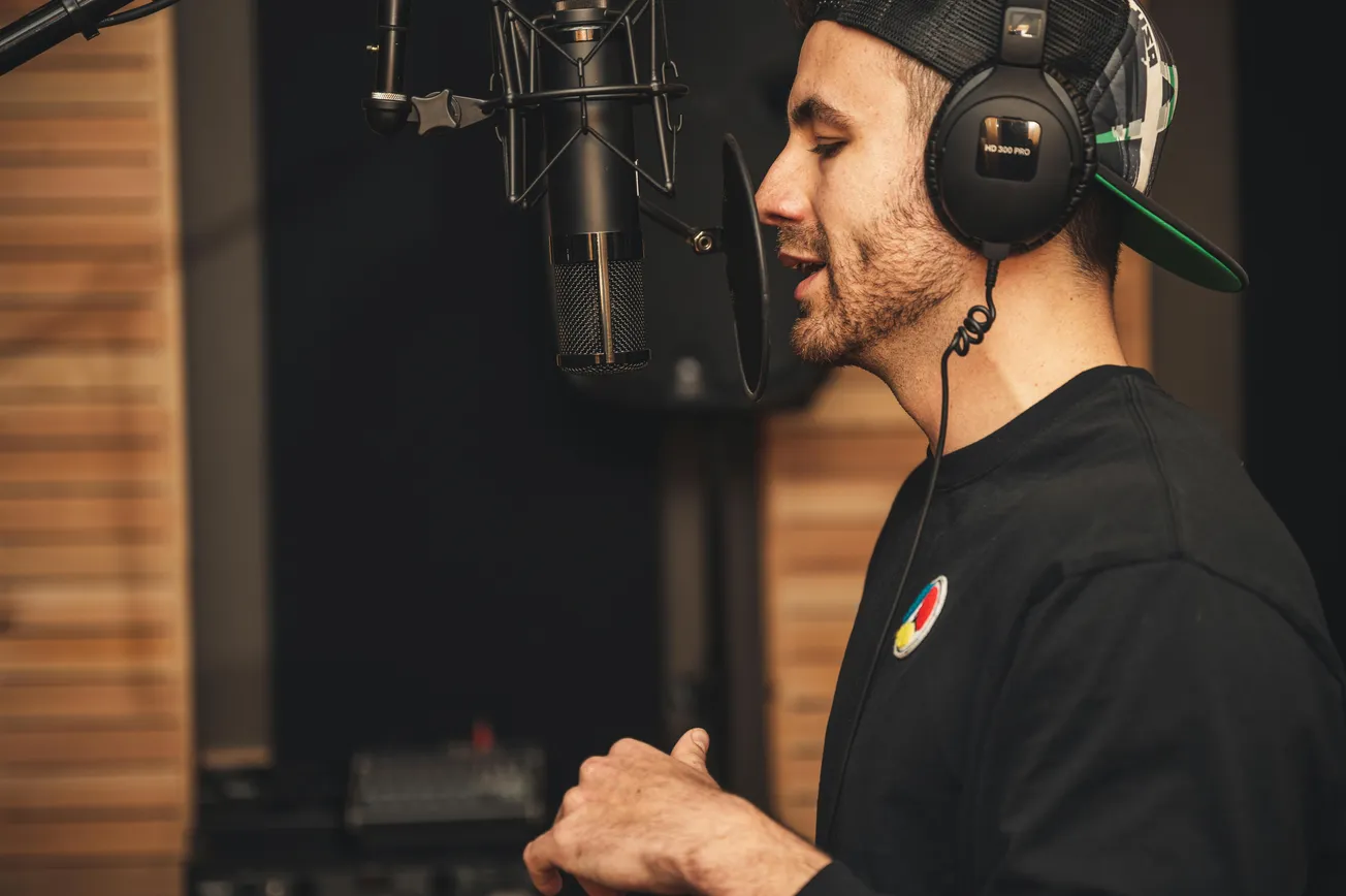 A man in a recording studio sings into a microphone, wearing large headphones and a black cap. The setting is warm and focused on music creation.