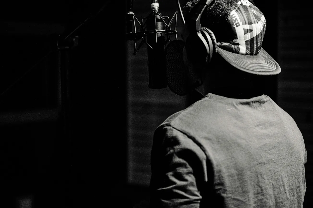 Black and white image of a person wearing a cap and headphones, facing a microphone in a recording studio. Dim lighting creates a focused, intimate mood.