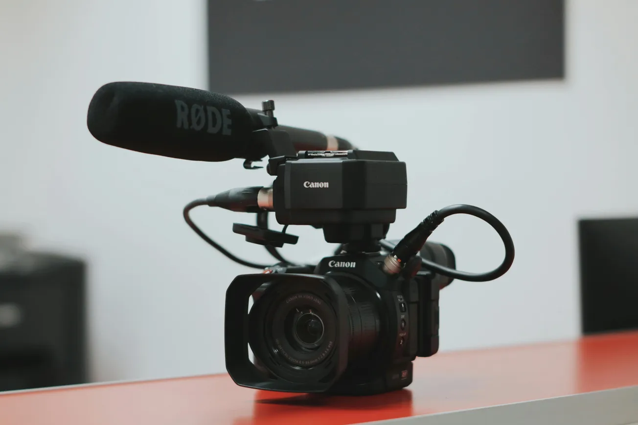 Close-up of a Canon video camera with a Rode microphone attached, placed on a red surface. The setting appears to be a professional studio environment.