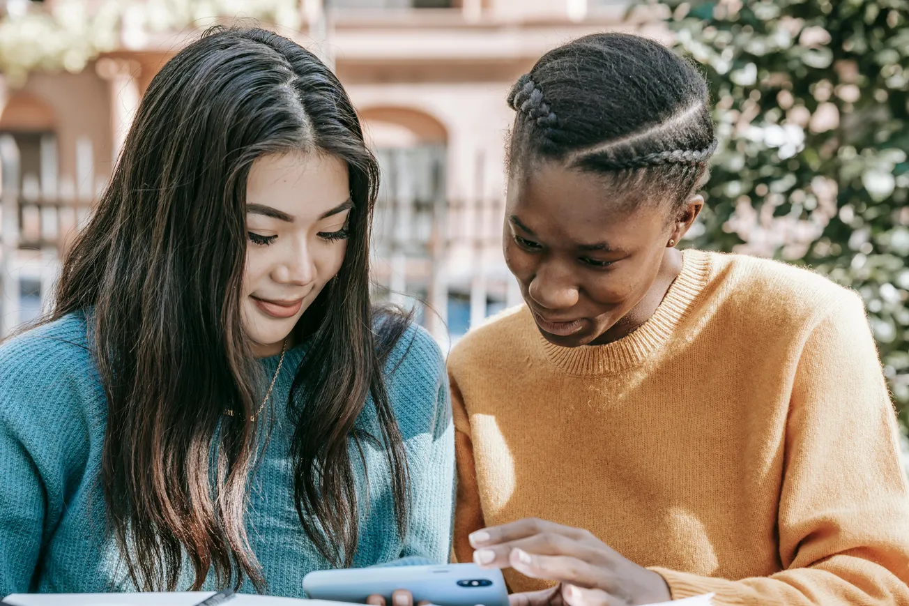 Two young women, one in a blue sweater and the other in orange, smile while looking at a smartphone outdoors. The scene conveys warmth and friendship.