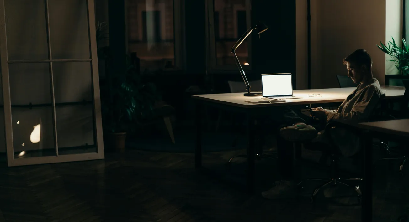 A person sits in a dimly lit room at a large desk with an open laptop, illuminated only by its screen. The atmosphere feels quiet and focused.