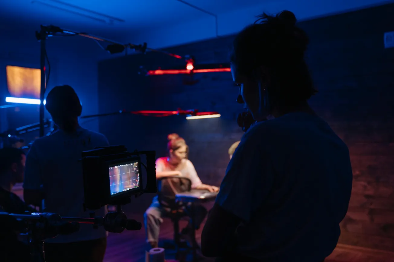 Behind-the-scenes of a film set with dim lighting and red-blue hues. A camera in the foreground captures two actors sitting at a table, focused and intense.
