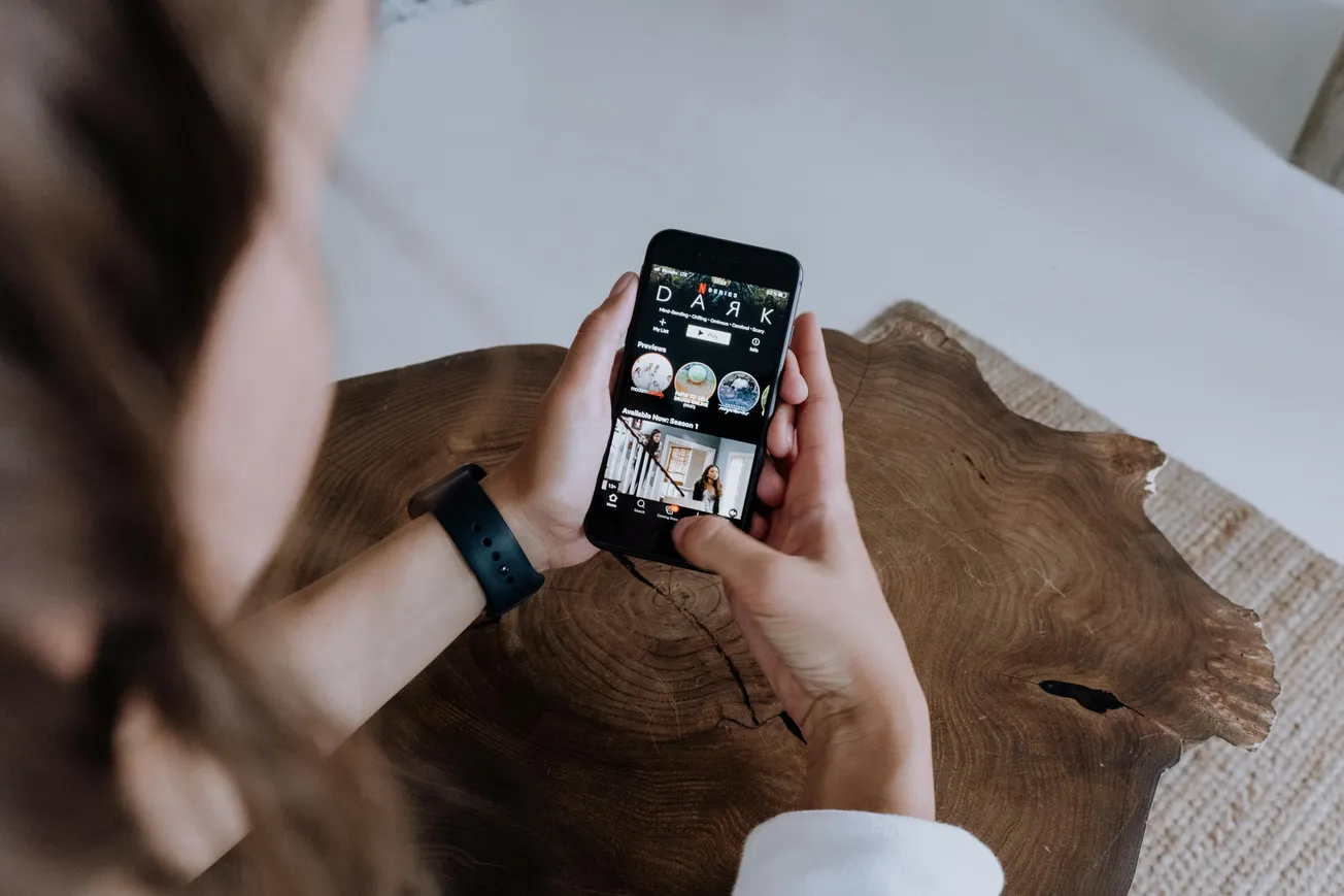A person holds a smartphone displaying a streaming service interface with thumbnails. They're seated at a wooden table, creating a casual, modern vibe.