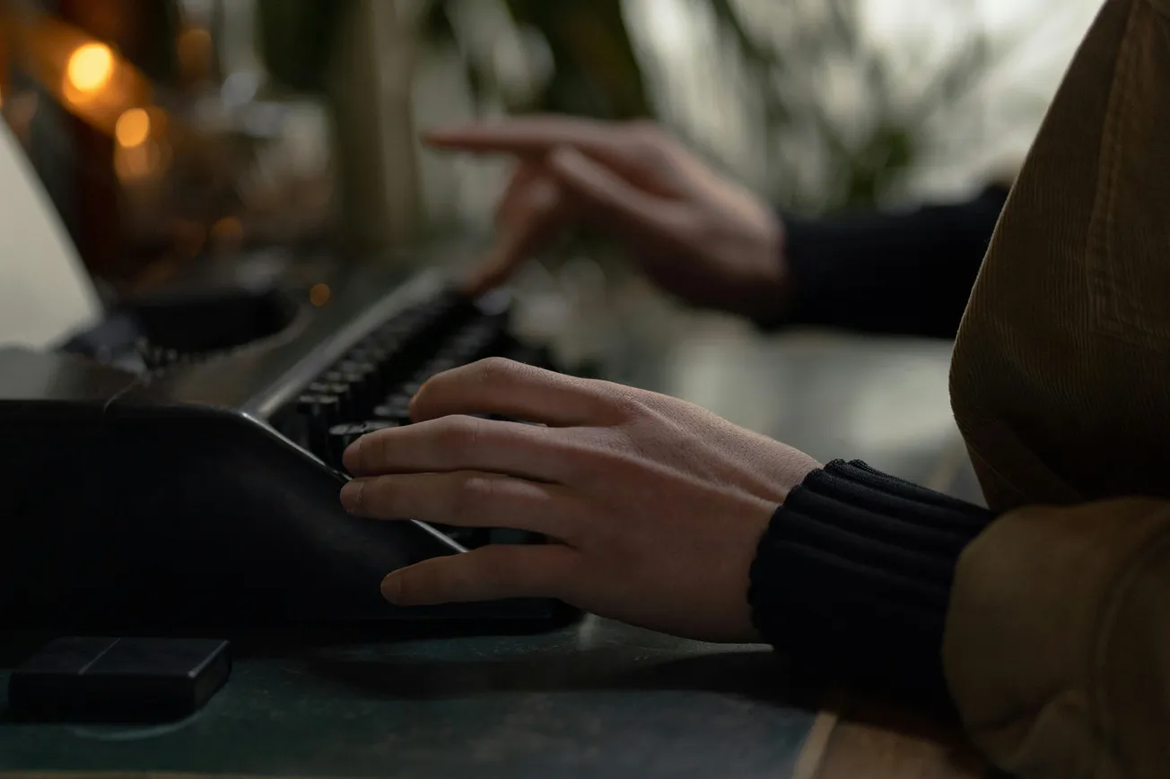 Hands typing on a vintage typewriter in dim light, creating a focused and nostalgic atmosphere. Blurred plants and warm tones in the background.