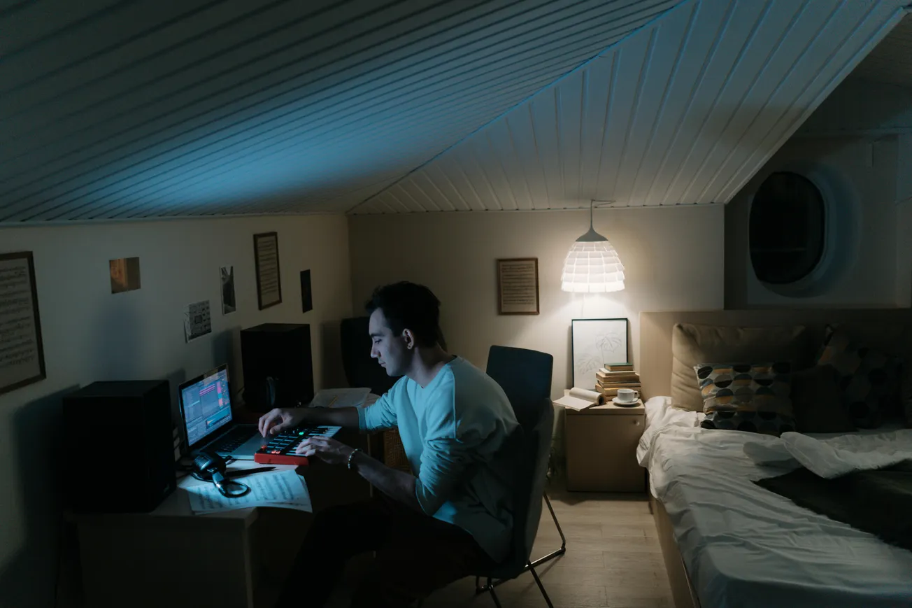 A person works intently on a computer in a dimly lit attic room, illuminated by a small desk lamp. The cozy space includes a bed and decorative items.