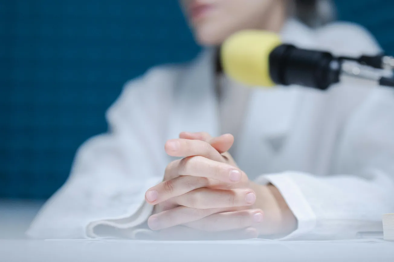 A person in a white coat sits at a table with hands clasped, next to a microphone with a yellow windscreen. The blue background adds a calm tone.