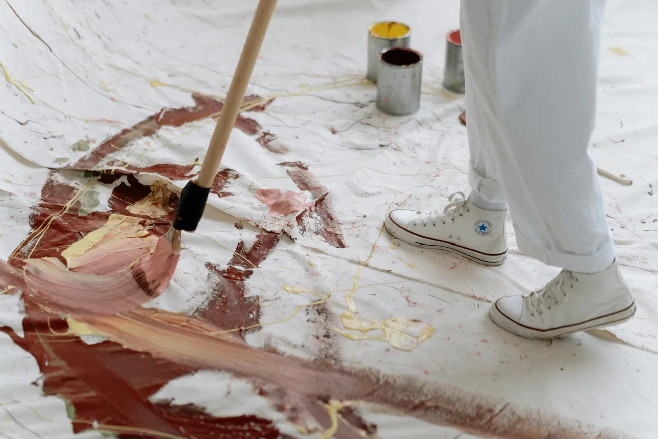 Person in white sneakers and pants painting a large canvas on the floor with abstract brown and yellow strokes, using a long brush.