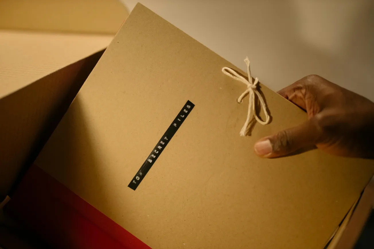 A hand holds a brown envelope labeled "Top Secret Files," tied with string, conveying intrigue. The envelope is partially inside a cardboard box.