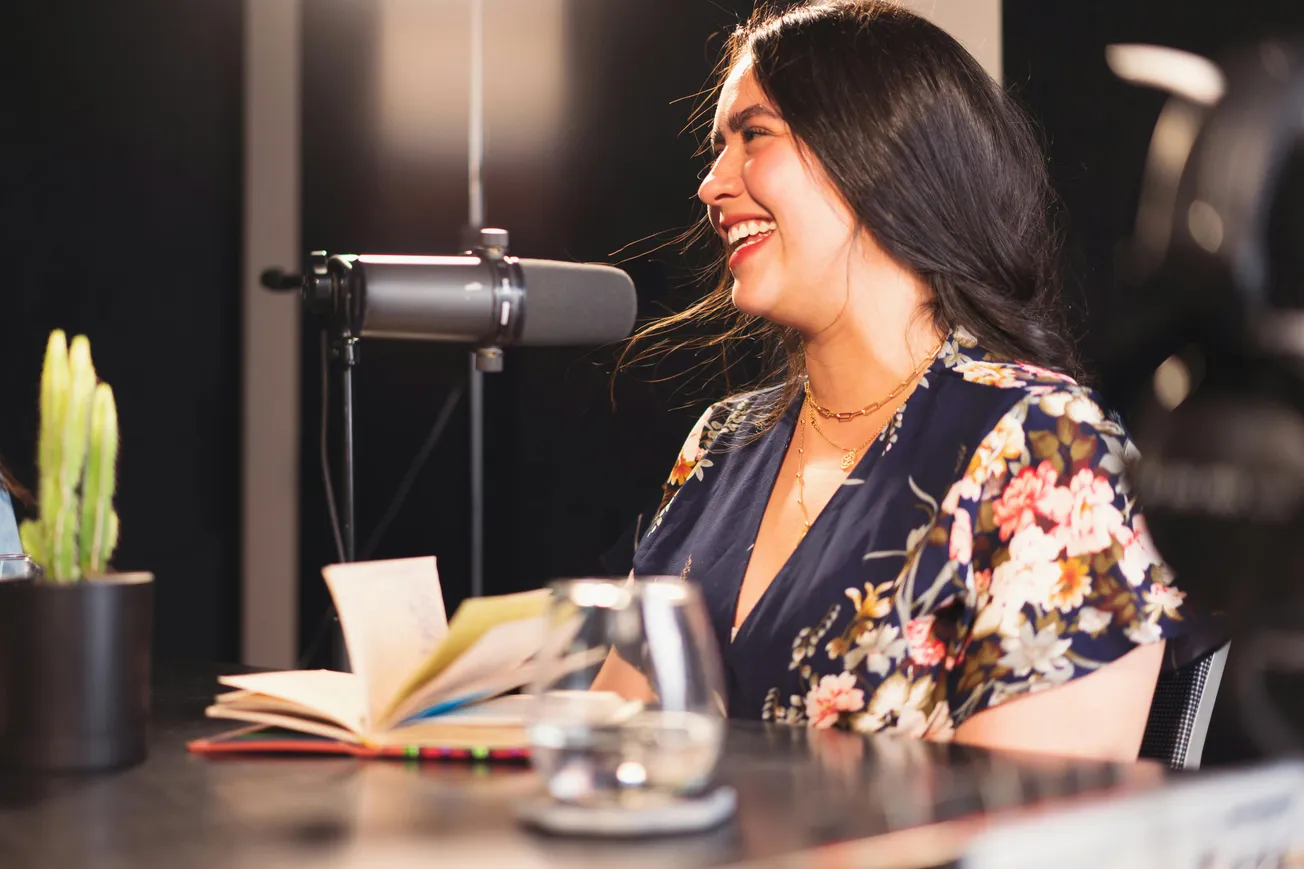 Woman with long dark hair in a floral dress, smiling while speaking into a microphone. A notebook and a glass are in front of her. Podcast setting.
