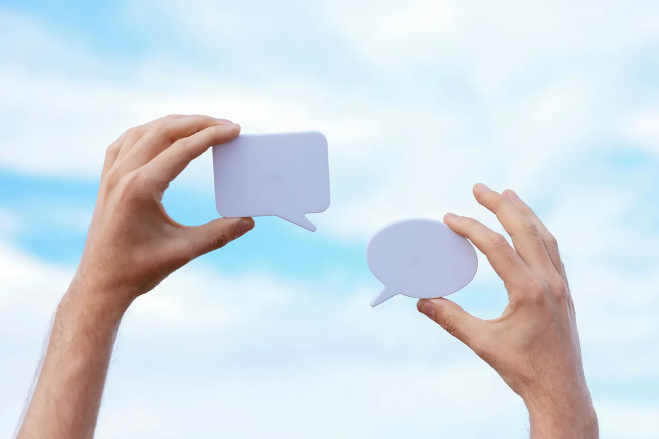 Hands holding two blank speech bubble cutouts against a bright, cloudy sky, conveying communication and conversation concepts.