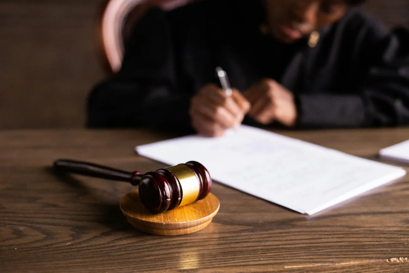 A wooden gavel rests on a desk in the foreground. In the background, a person in a black robe writes on papers, conveying a legal or judicial setting.