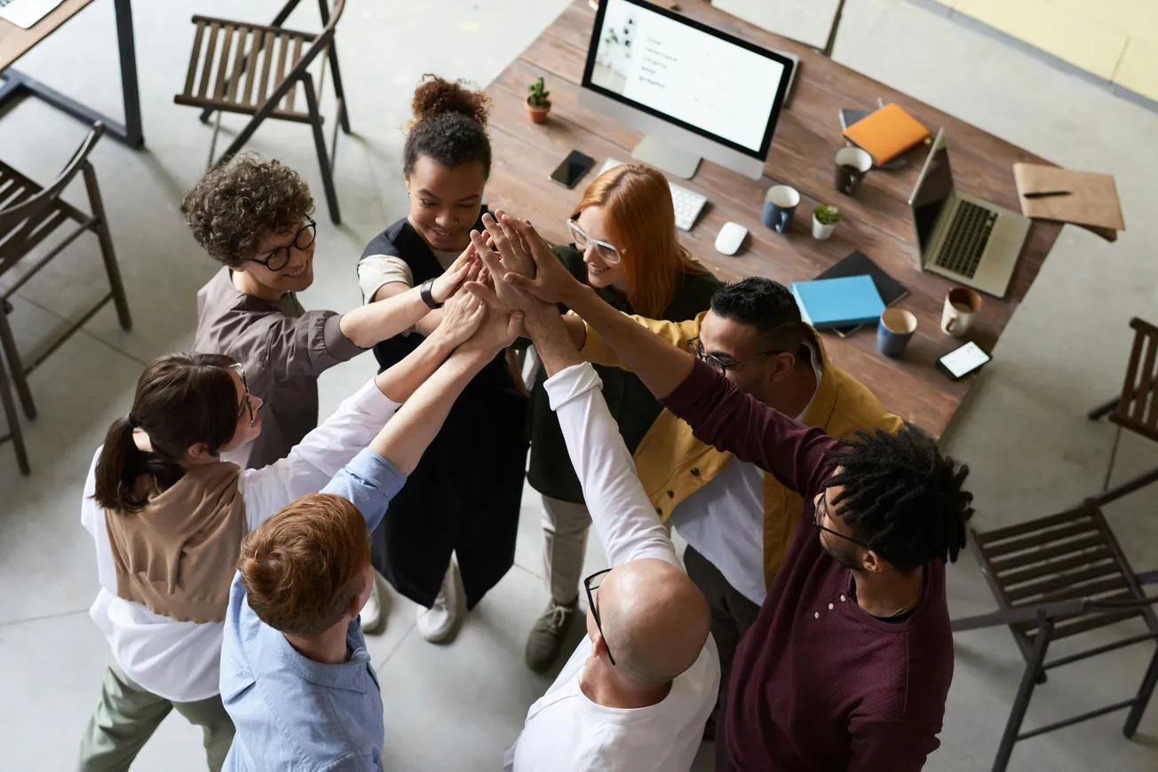 A diverse group of people stands in a circle, reaching in for a collective high-five, smiling and expressing teamwork in a collaborative office space.