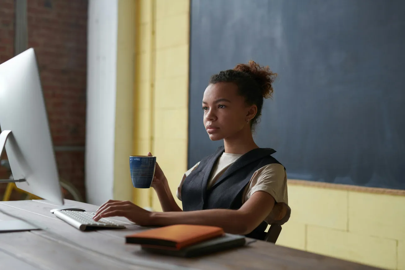 A focused woman sits at a wooden desk with a computer, holding a mug. A notepad lies nearby. The background has a chalkboard and yellow wall.