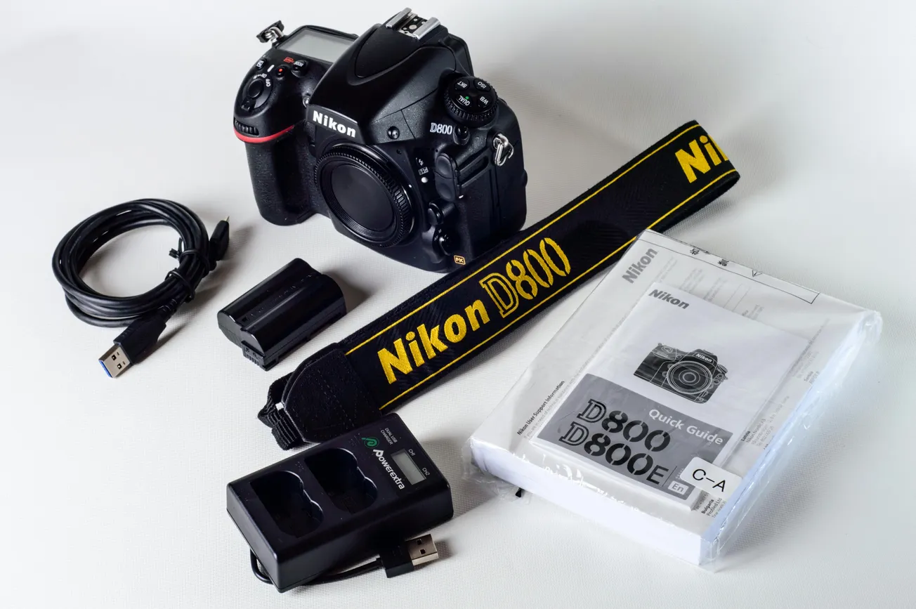 Nikon D800 camera kit with body, USB cable, battery, charger, neck strap, and instruction manuals. Items are neatly arranged on a white surface.