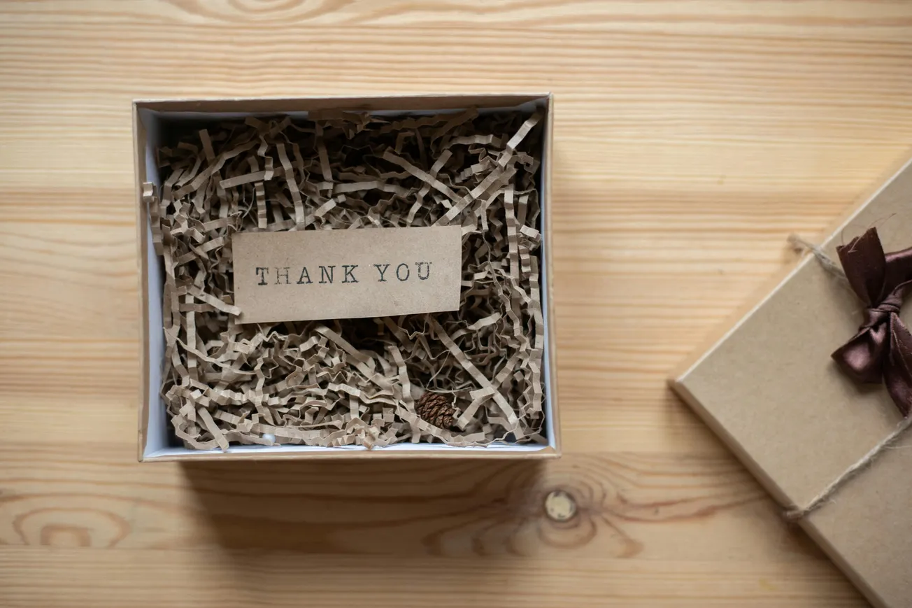 A gift box filled with shredded paper holds a small card reading "Thank You." The box, placed on a wood surface, conveys appreciation and warmth.