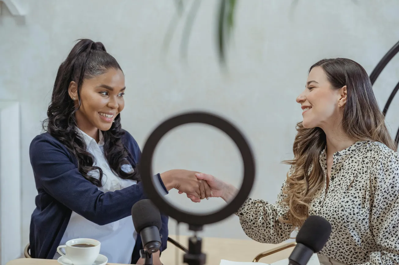 Two women smiling and shaking hands across a table. A ring light and microphones are visible, suggesting a friendly podcast or interview setting.