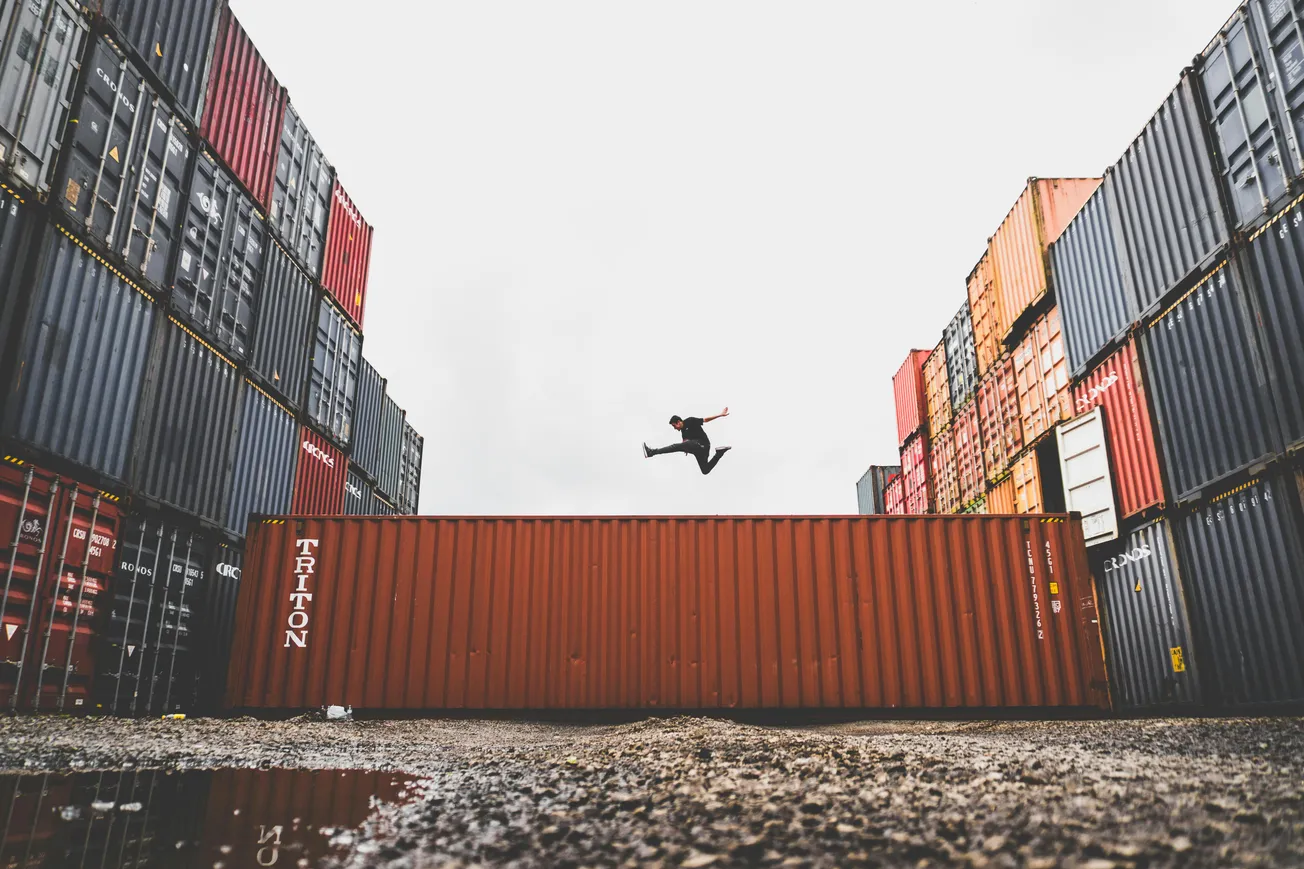 Person in mid-air, performing a dynamic jump between stacked rows of colorful shipping containers against an overcast sky, conveying energy and freedom.