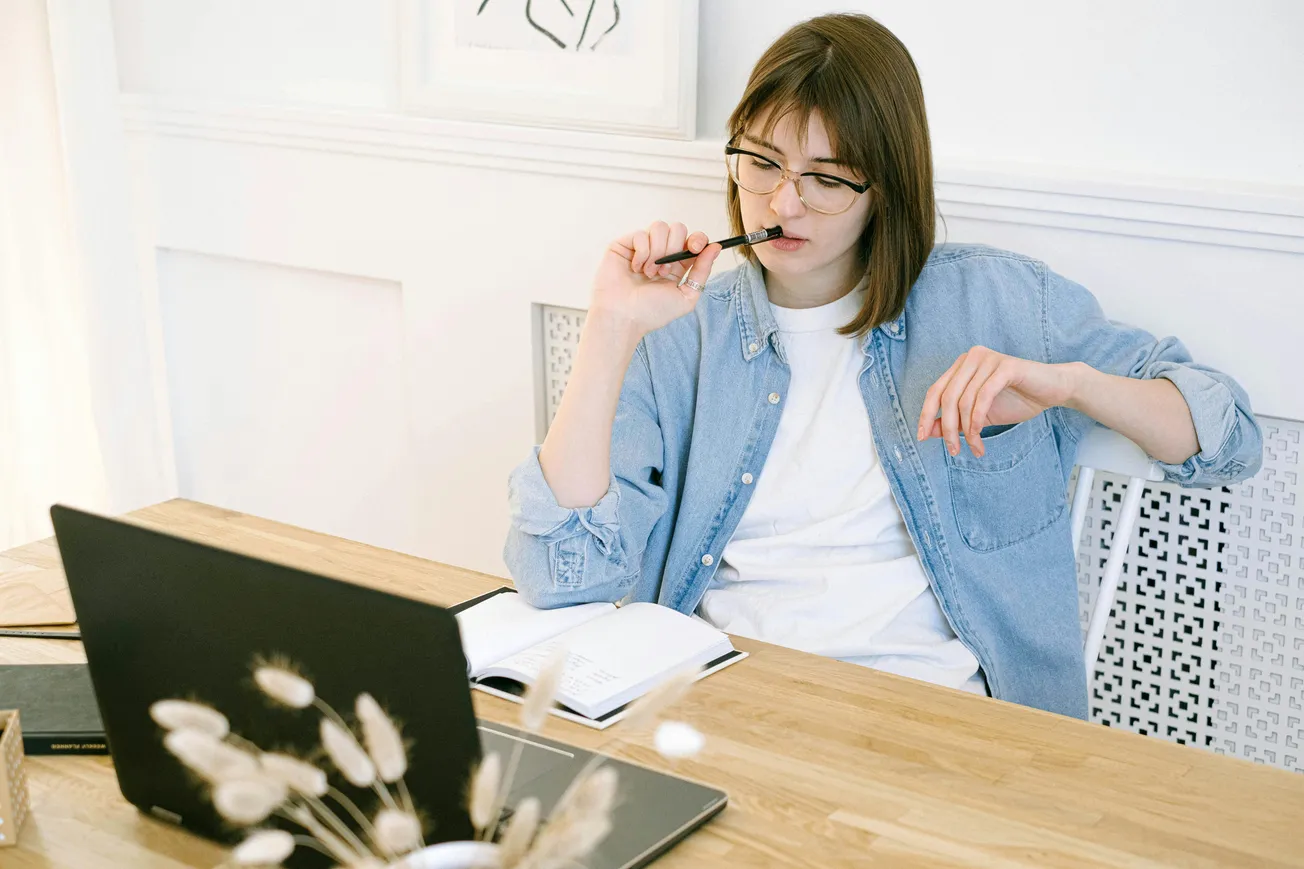 A woman wearing glasses and a denim shirt sits pensively at a desk with an open notebook and laptop. She holds a pen to her lips, conveying deep thought.