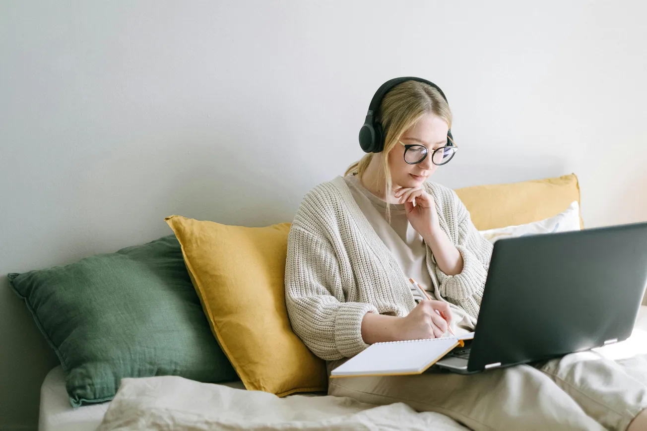 A woman with headphones sits on a bed, focused on a laptop, and taking notes in a notebook. She is surrounded by gray and yellow pillows, conveying a calm, productive atmosphere.