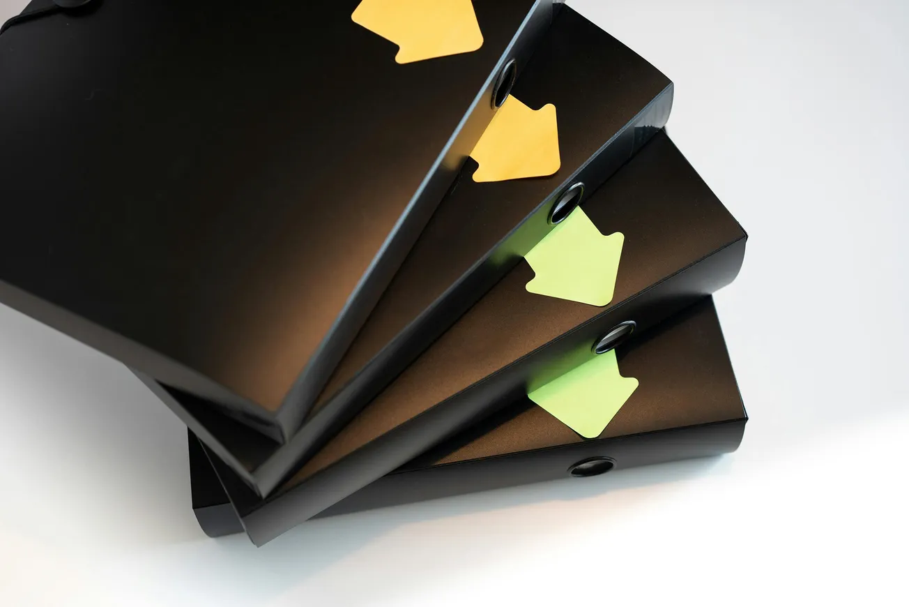 A stack of four black file folders with circular holes, each marked with colorful arrow-shaped sticky notes in yellow and green, lying on a white surface.
