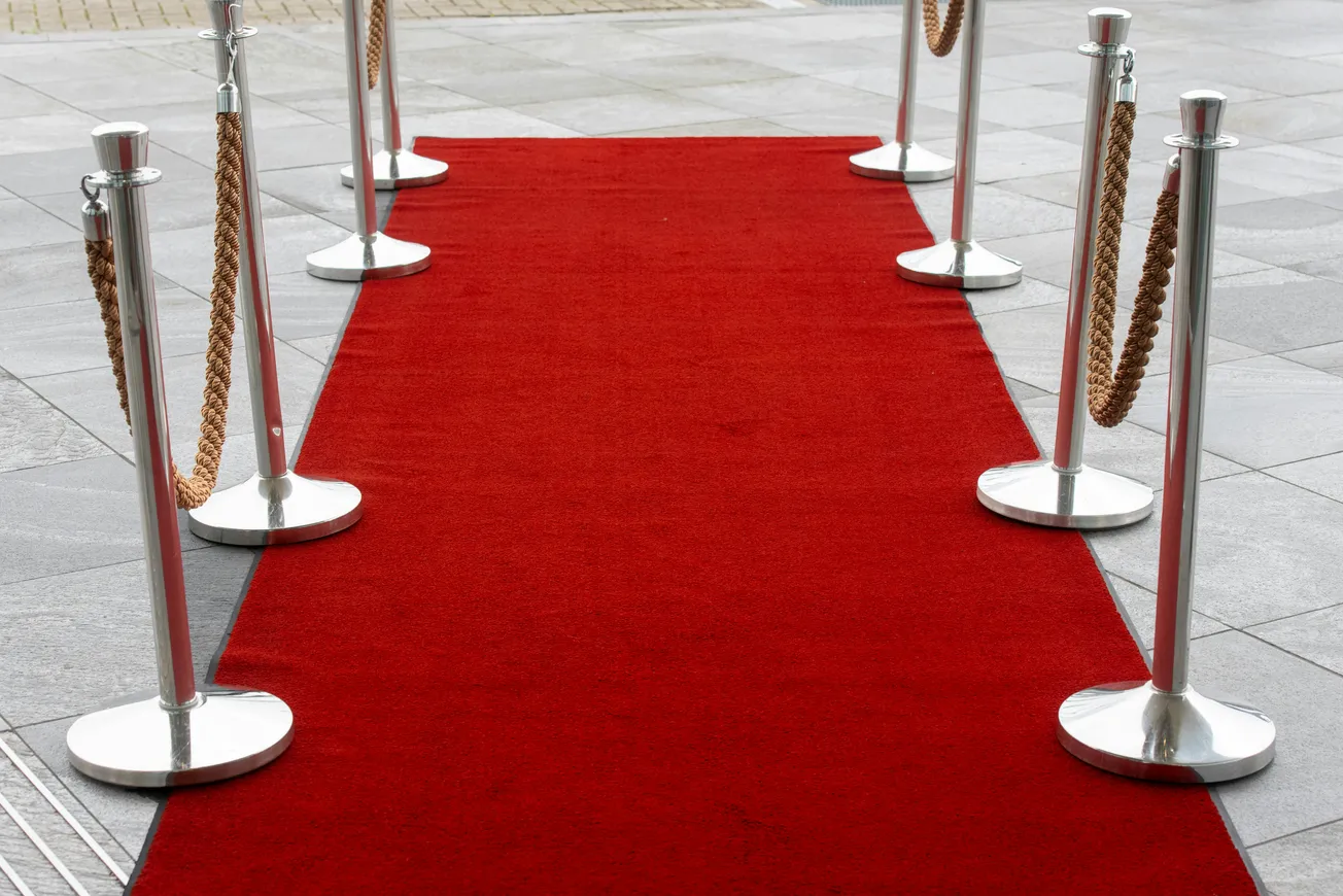 Red carpet lined with silver stanchions and velvet ropes on a gray tiled floor. The setup conveys a sense of elegance and exclusivity.