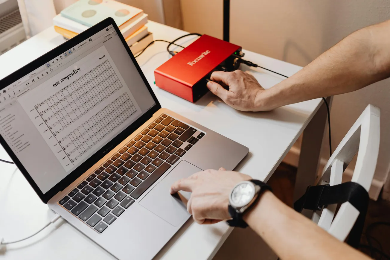 A person works on a music composition on a laptop with sheet music displayed. Their hand adjusts a red audio interface on a white desk, conveying creativity.