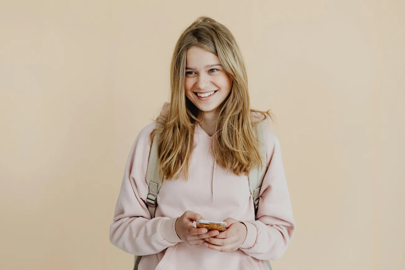 Smiling girl in a pink hoodie holds a smartphone against a beige background. She wears a backpack and exudes a cheerful, relaxed vibe.