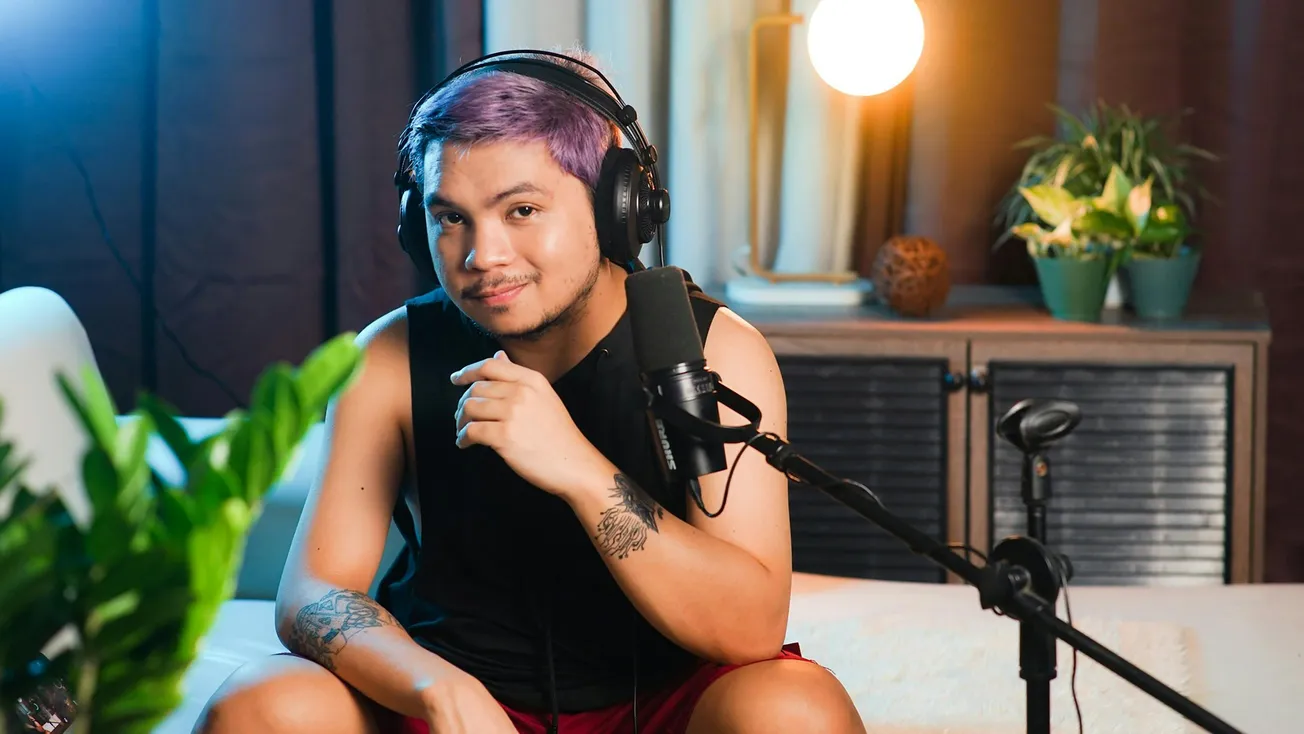 A person with purple hair and tattoos sits on a couch, wearing headphones and speaking into a microphone. The room is warmly lit with plants nearby.