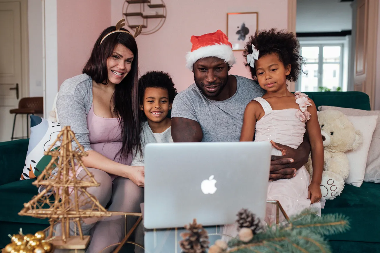 Family on sofa, parents and two kids, smiling while video chatting on a laptop. A cozy Christmas setting with festive decorations and a teddy bear.