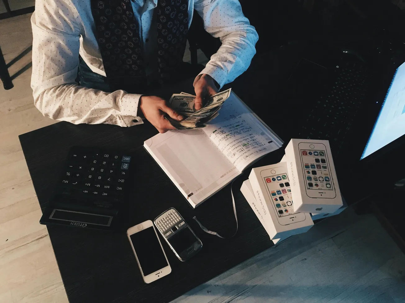 A person in a patterned shirt counts money at a desk with a notebook, stacked phone boxes, calculator, and phones. The setting conveys business activity.