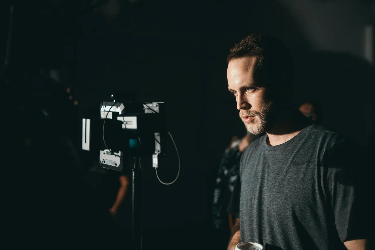 Man in a dimly lit room stands near camera equipment, looking focused. He wears a dark T-shirt, holding a cup, conveying a professional atmosphere.