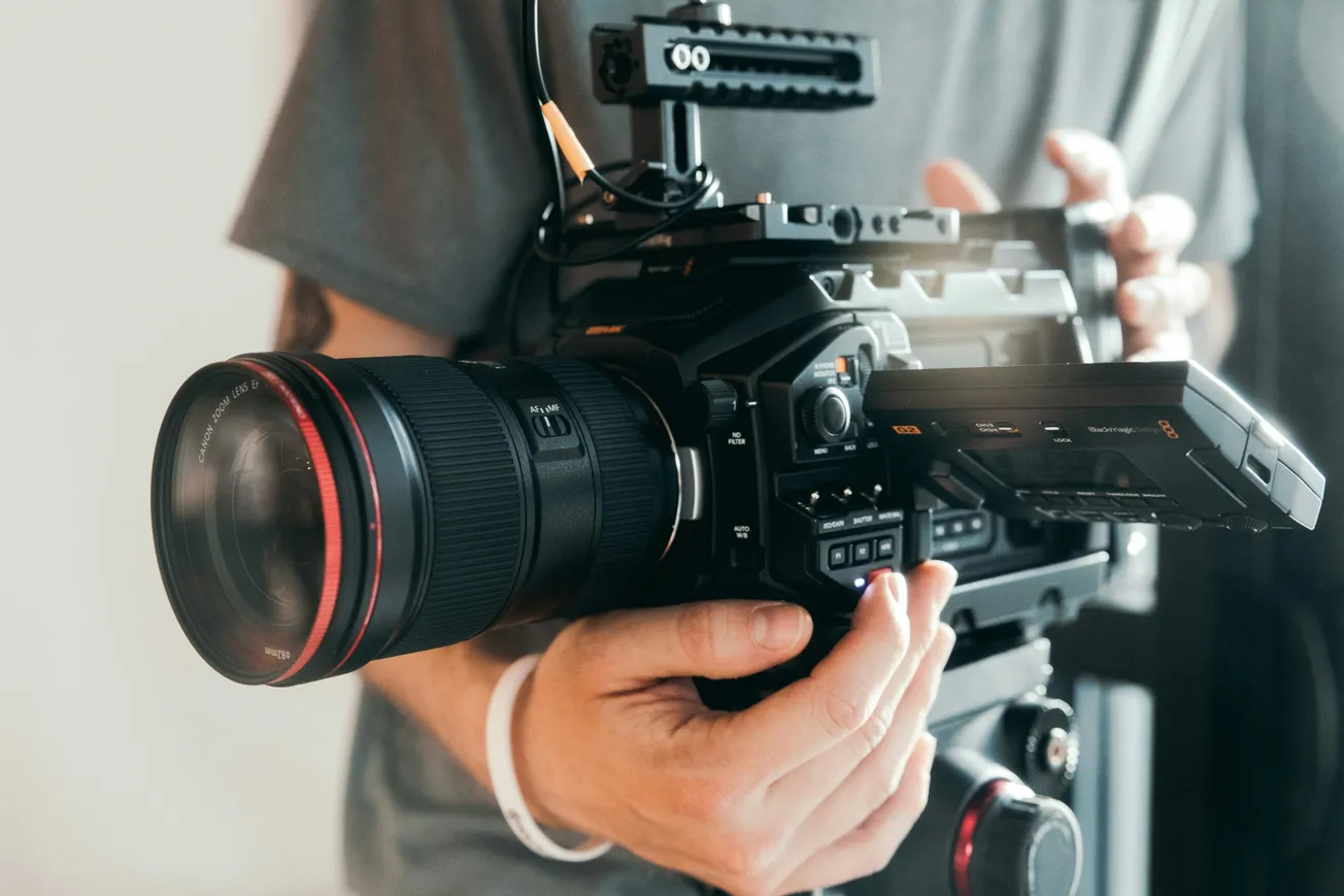 Close-up of hands holding a professional video camera with a large lens and side screen. The scene conveys a sense of focus and creativity.